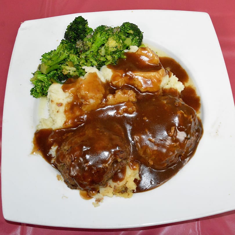 Two Salisbury steaks covered in gravy, with mashed potatoes and steamed broccoli on a white plate.